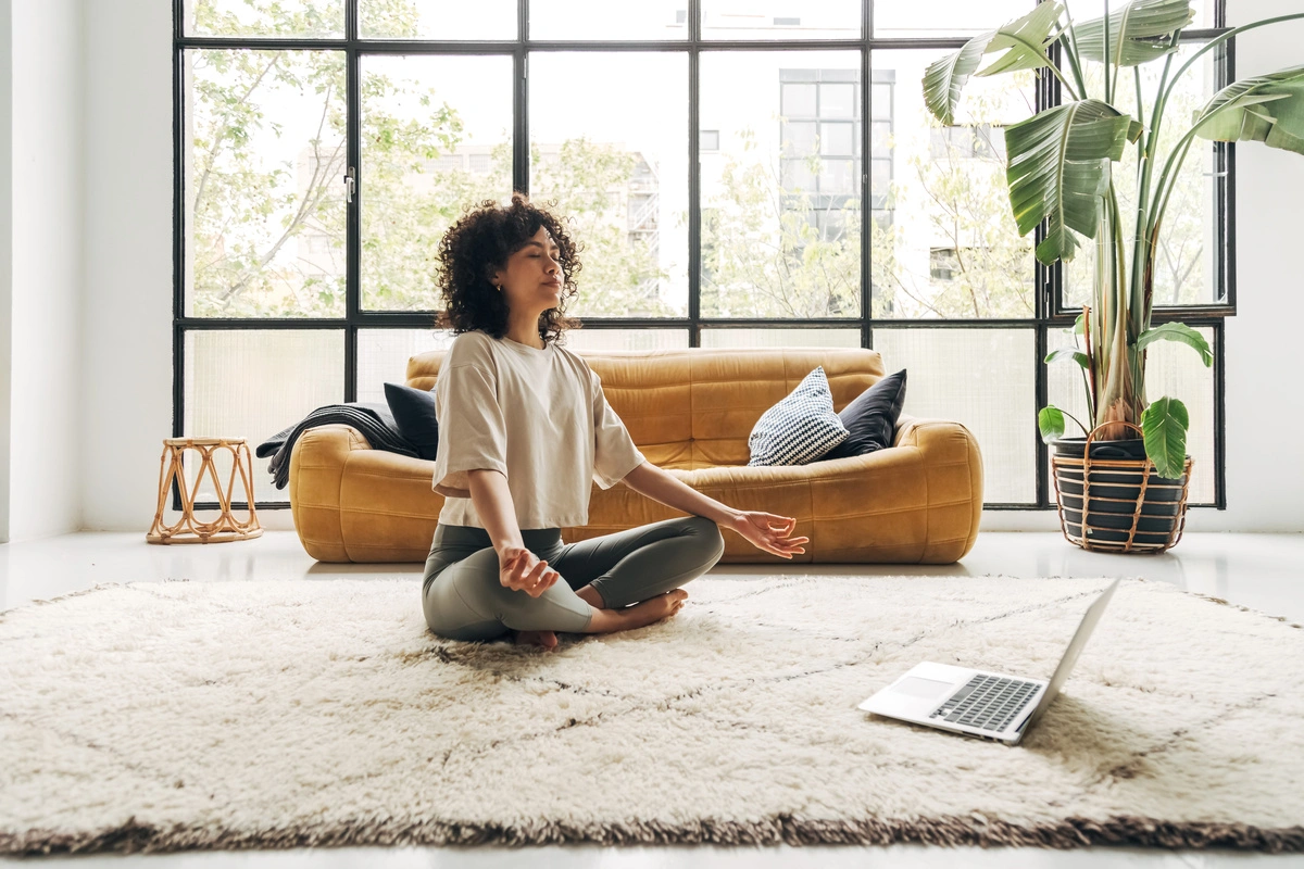 Jeune femme méditant en ligne dans son salon, assise en tailleur sur un tapis de yoga, avec un ordinateur portable posé sur le sol.